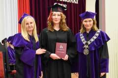 Absolwentka w czarnej todze i birecie stoi między dwiema kobietami w fioletowych togach; trzyma czerwony dyplom podczas uroczystości w auli