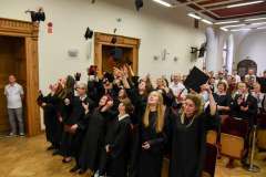 Absolwenci w czarnych togach podrzucają birety w górę podczas uroczystości; w tle stoją i klaszczą zaproszeni goście