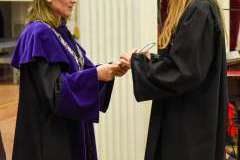 Kobieta w fioletowej todze i birecie wręcza dokument studentce w czarnej todze i birecie podczas uroczystości absolutoryjnej w auli