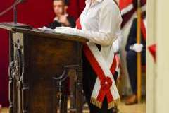 Studentka w birecie i z biało-czerwoną szarfą przemawia przy mównicy podczas uroczystości absolutoryjnej; w tle widoczne flagi i czerwone kotary.