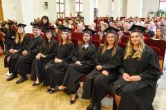 Grupa absolwentów w czarnych togach i biretach siedzi w pierwszym rzędzie auli podczas uroczystości, w tle widoczni goście