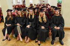 Grupa absolwentów w czarnych togach i biretach siedzi w pierwszym rzędzie auli podczas uroczystości, w tle widoczni goście