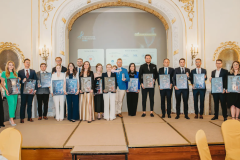 Grupa stojących ludzi, stojących w półkolu w eleganckich wnętrzach budynku, trzymająca w rękach nagrody