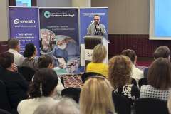 Zdjęcie przedstawia konferencję medyczną, prelegent mówi przez mikrofon i pokazuje slajd, uczestnicy siedzą i słuchają prezentacji.