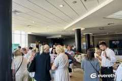 Uczestnicy konferencji podczas przerwy, ludzie stoją przy stołach z poczęstunkiem i rozmawiają w grupach w jasnym holu.