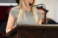 Młoda kobieta stoi przed mównicą i przemawia przez mikrofon. Atmosfera jest uroczysta.