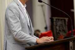 Mężczyzna stoi przed mównicą i przemawia przez mikrofon. Atmosfera jest uroczysta.