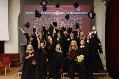 Grupa studentów ubranych w togi rzuca biretki w górę. Za nimi tablo całego rocznika