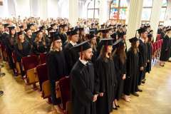 Grupa studentów w togach i biretkach stojących w auli uczelni. Atmosfera jest uroczysta.