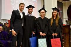 Uśmiechnieci studenci ubrani w togach i biretkach trzymają w ręku papierowe torebki 