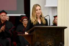 Młoda kobieta stoi przed mównicą i przemawia przez mikrofon. Atmosfera jest uroczysta.