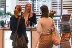 dwie kobiety stoją przy stoisku promocyjnym i słuchają trzeciej kobiety