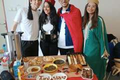 Cztery młode uśmiechnięte osoby w narodowych strojach stoją pred stołem z jedzeniem na talerzykach