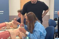 Grupa młodych osób w ubraniu medycznym w sali warsztatowej. na stole leżą fantomy do intubacji