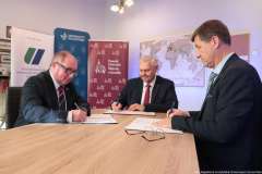 Trzej mężczyźni w garniturach podpisują dokumenty przy stole; w tle widoczne ścianki z logo ZUT, Uniwersytetu Szczecińskiego i PUM