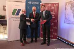 Trzej mężczyźni w garniturach stoją przed ściankami z logo Uniwersytetu Szczecińskiego, ZUT i PUM, ściskają sobie dłonie i trzymają podpisane dokument