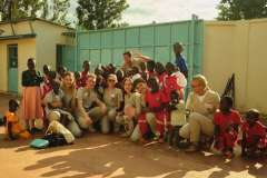 Medycy w uniformach lekarskich wśród grupy dzieci. Niektórzy stoją, niektórzy kucają. Wszyscy są uśmiechnięci