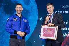 dwóch mężczyzn na scenie, jeden mówi przez mikrofon i trzyma dylom w ręku, drugi uśmiecha się