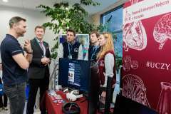 rozmowa dwóch mężczyzn z młodymi ludźmi przy stoisku prezentującym ich pracę, w tle ścianka promocyjna