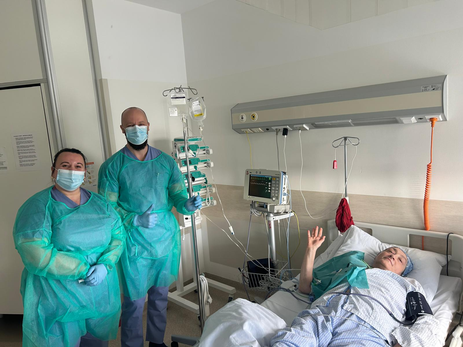 Kobieta i mężczyzna ubrani w stroje medyczne oraz maski na ustach stoją w sali chorych. Na łóżku szpitalnym lezy kobieta która macha ręką